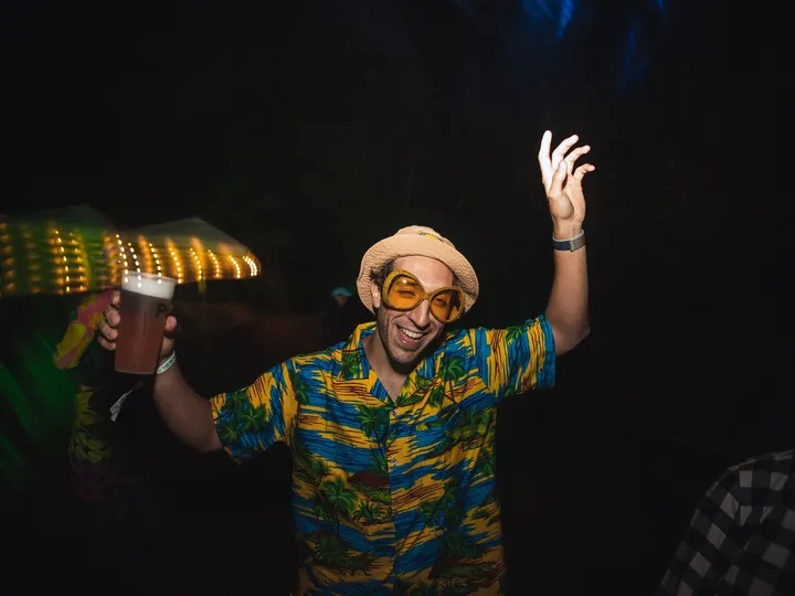 Un chico con camisa hawaiana, sombrero y gafas amarillas levanta el brazo en la fiesta