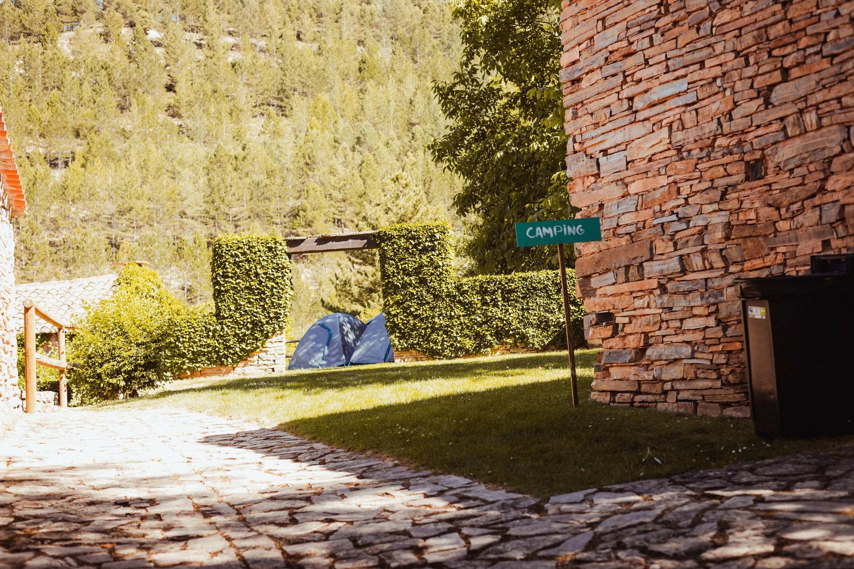 Calle empedrada junto a una casa de piedra con el cartel de «Camping» y una tienda montada al fondo, entre setos