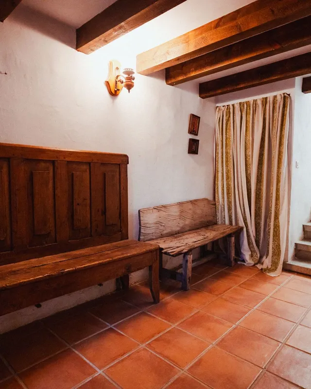 Recibidor de una casa del pueblo con un banco de madera, suelo de barro y la escalera a las habitaciones