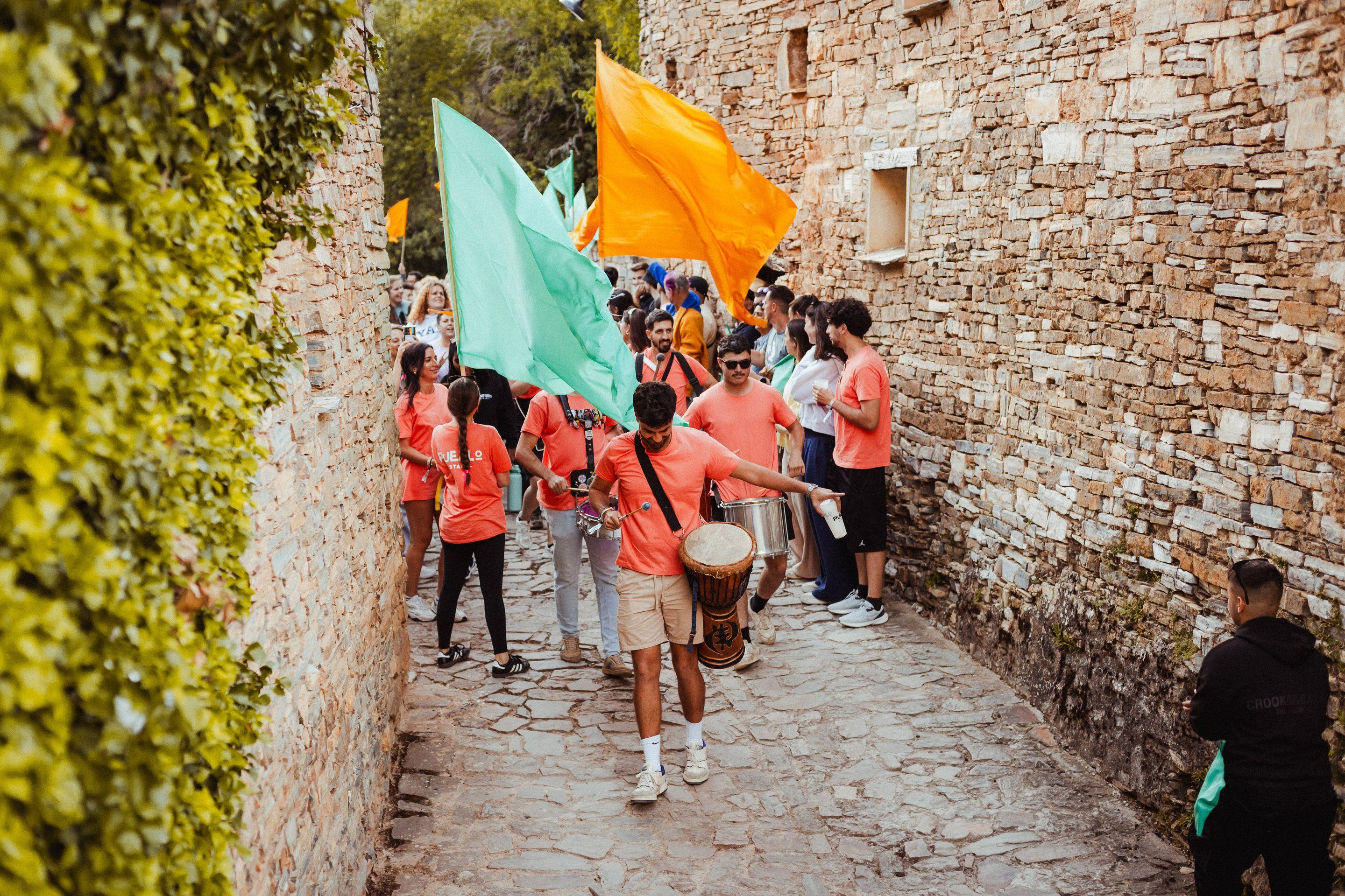 La charanga baja por una calle de piedra con las banderas turquesa y naranja de las familias