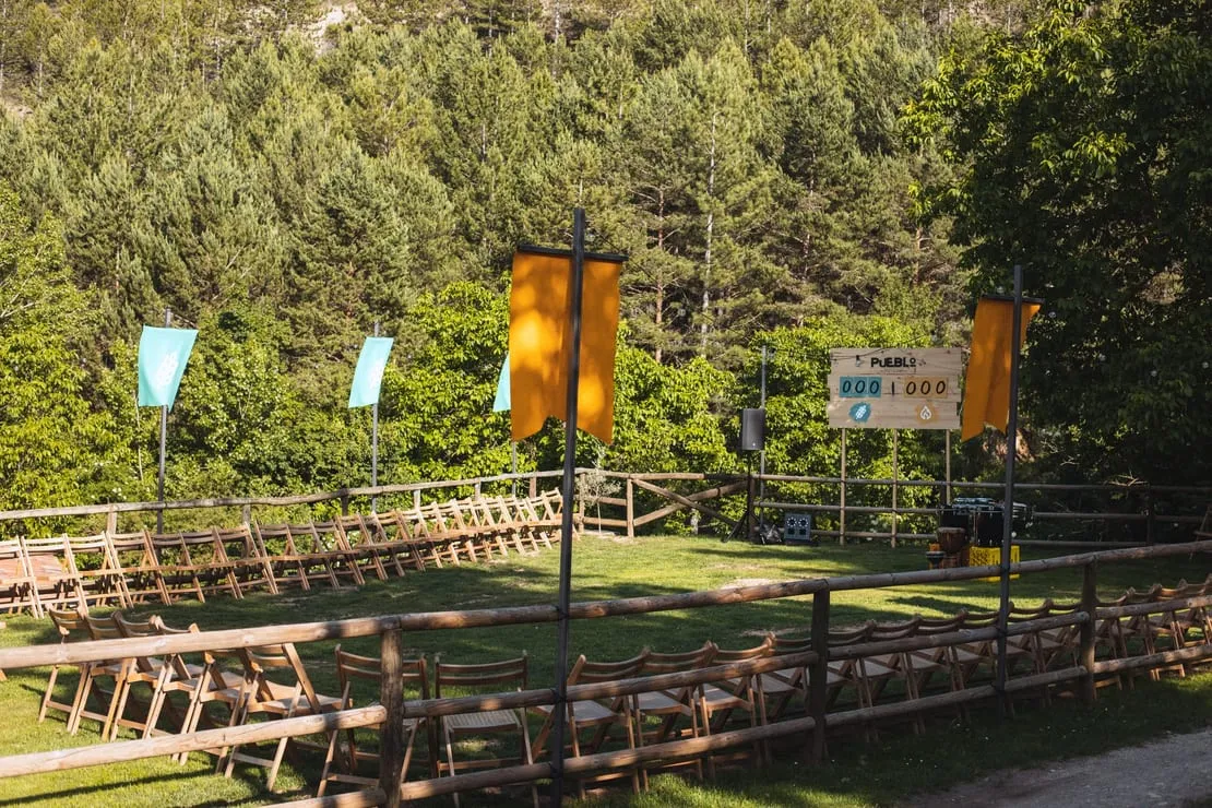 Sillas de madera organizadas en círculo en un campo rodeado de árboles. Banderas amarillas y azules y tabla de puntuación al fondo.