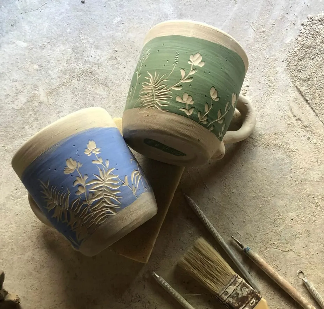 Dos tazas de cerámica decoradas a mano con motivos de plantas, junto a pinceles
