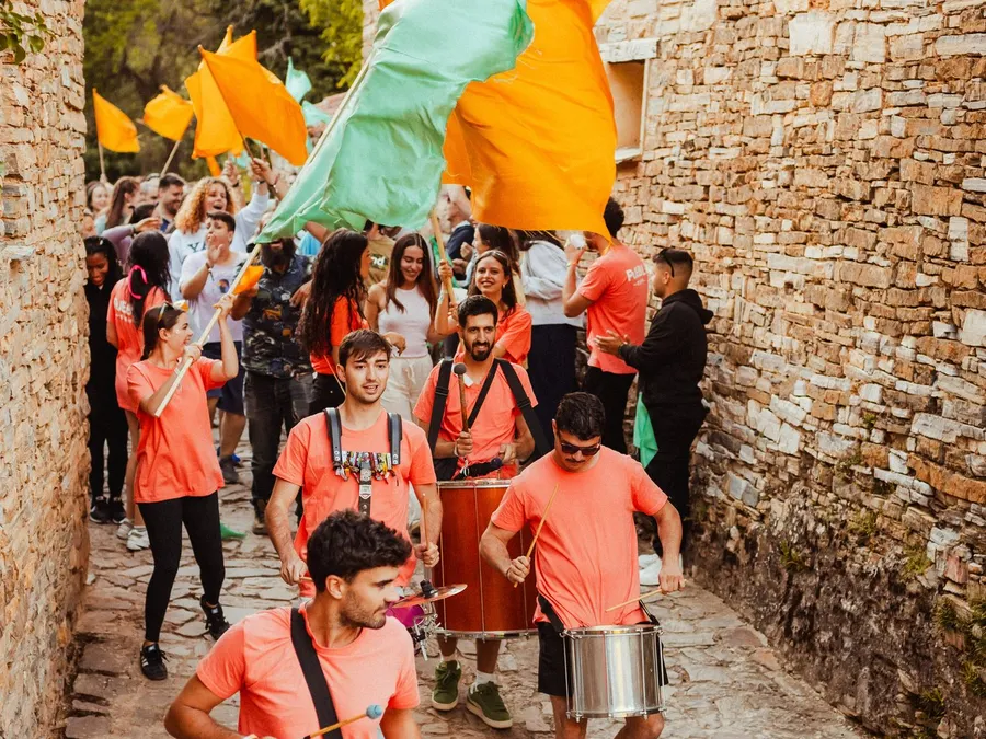 La batucada de PUEBLO, con camisetas naranjas y tambores, baja por una calle de piedra de Valdelavilla entre banderas naranjas y turquesas de las Familias