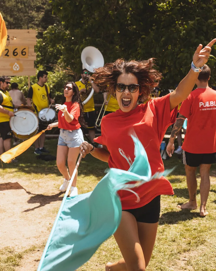 Una chica con camiseta roja de PUEBLO corre gritando y agita una bandera turquesa delante de la charanga