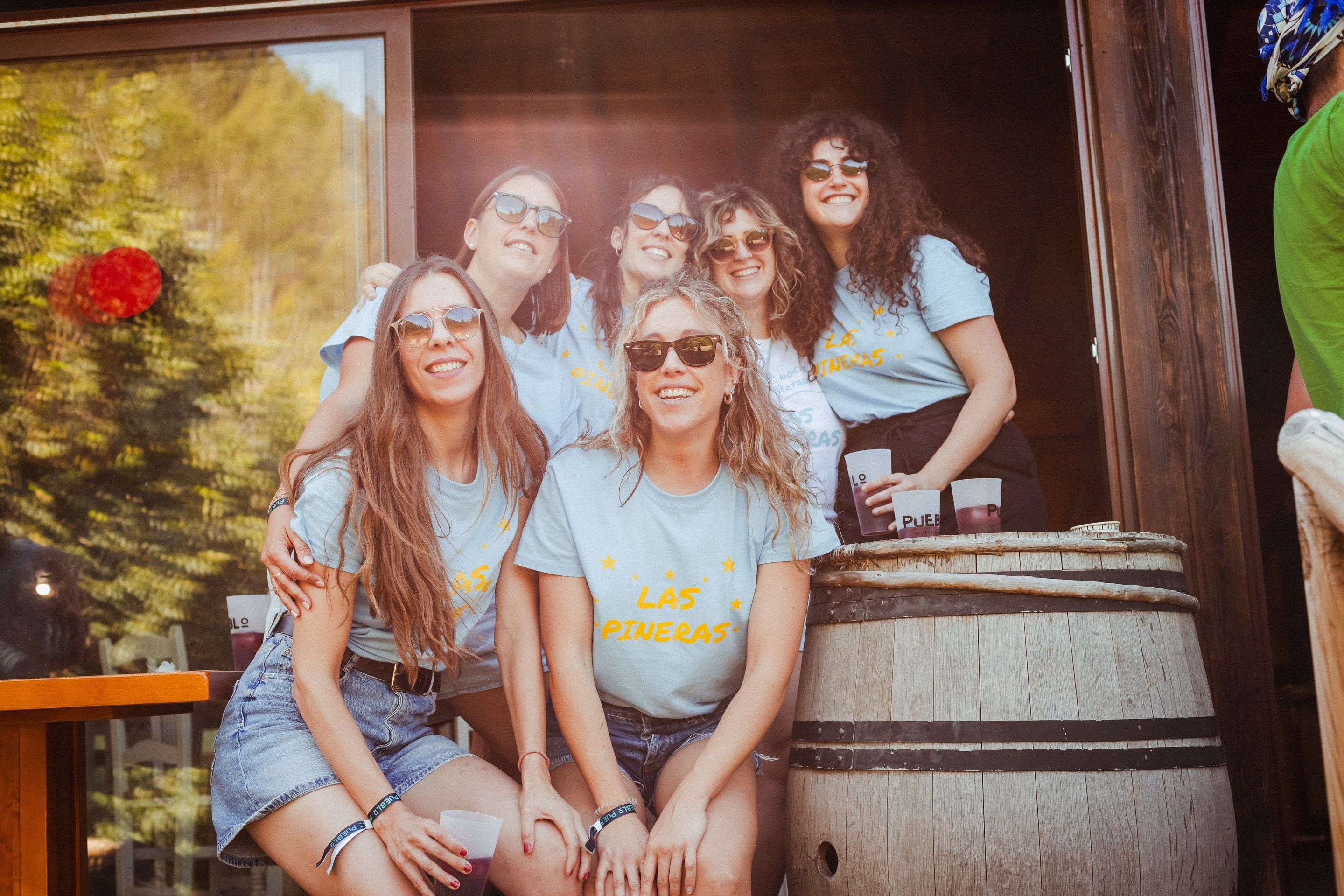 Un grupo de amigas con camisetas iguales y gafas de sol posa sonriente junto a un barril en el porche de PUEBLO Fest Camp