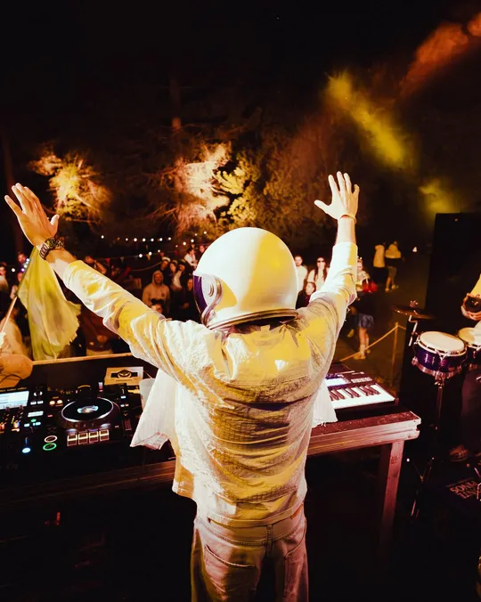 DJ con casco de astronauta y los brazos en alto ante el público en la fiesta