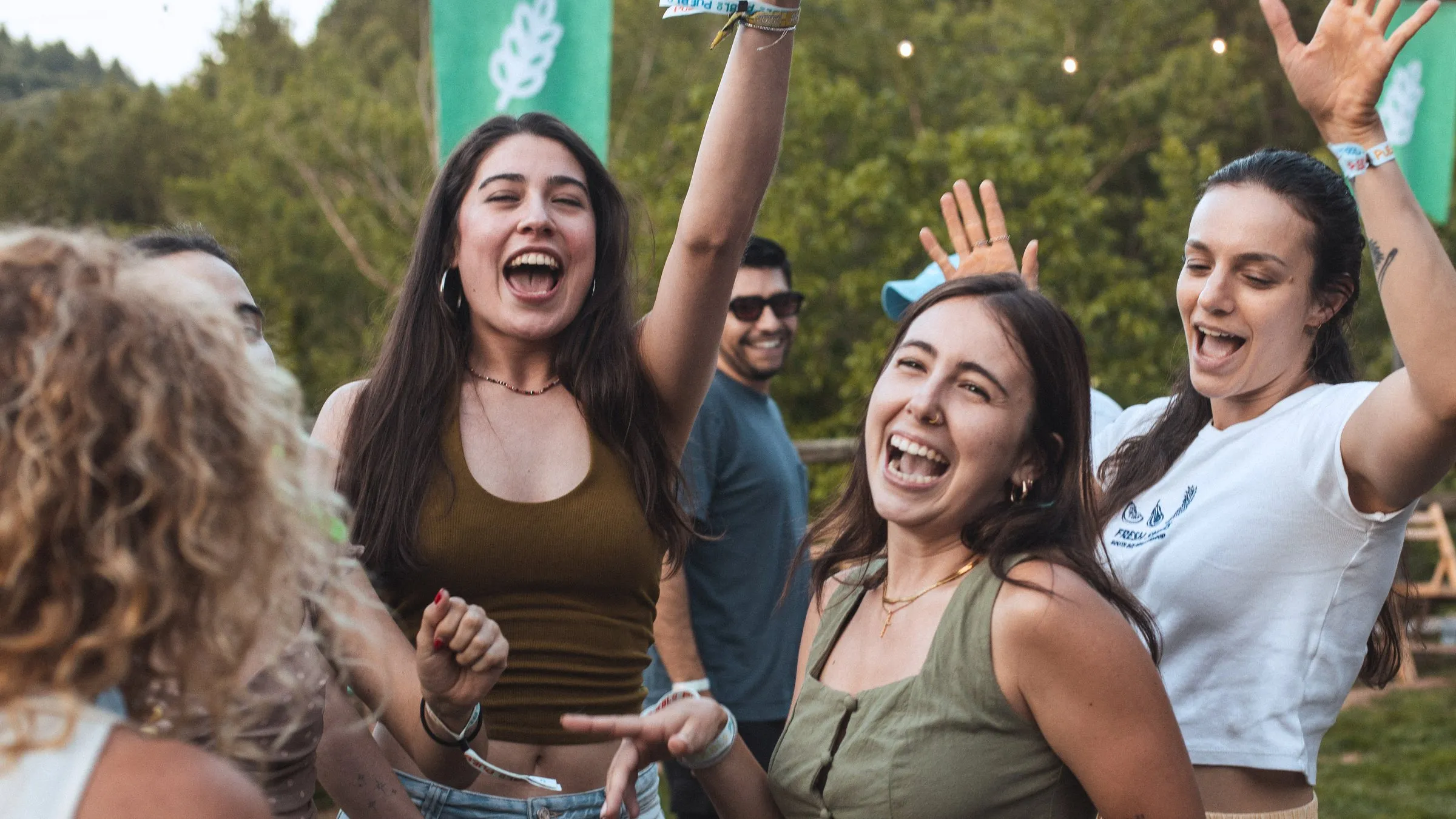 Unas amigas se ríen a carcajadas con los brazos en alto bajo la bandera turquesa en una tarde de PUEBLO 2025