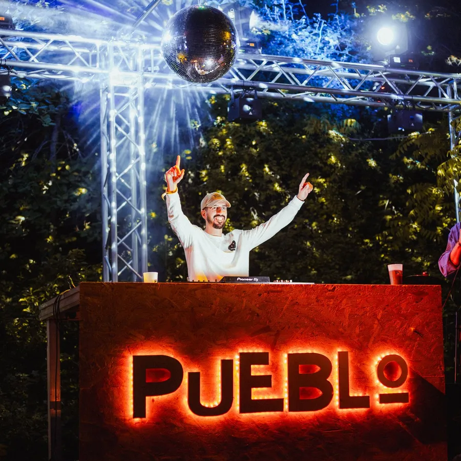 El DJ levanta los brazos en la cabina con el cartel luminoso de PUEBLO, bajo una bola de espejos