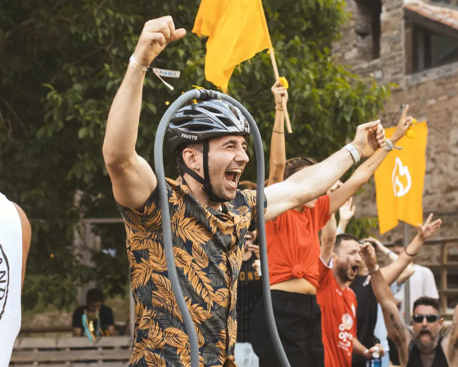 Un chico con casco de bici celebra a gritos con el puño en alto entre su equipo y las banderas naranjas