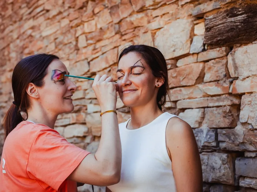 Una monitora pinta la cara a una chica en el taller de pintacaras, junto a un muro de piedra