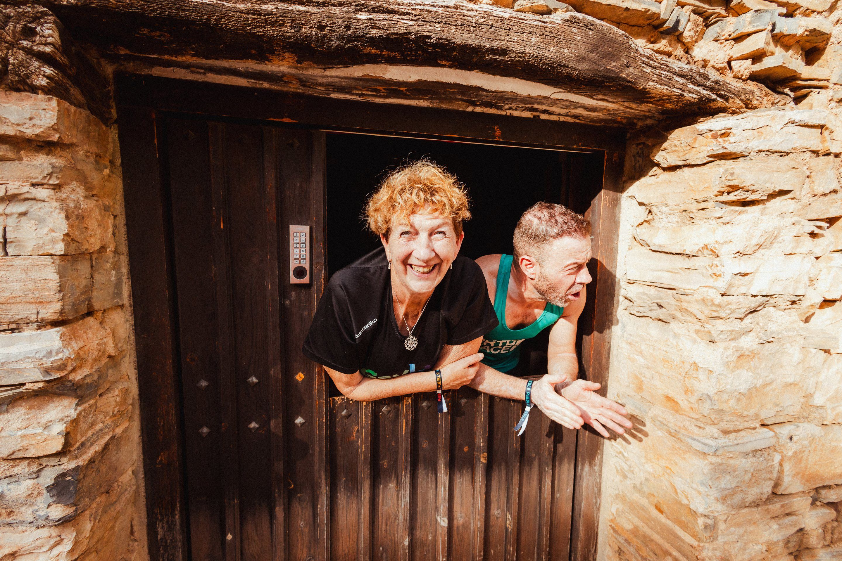 Dos personas se asoman sonriendo por la puerta partida de una casa de piedra